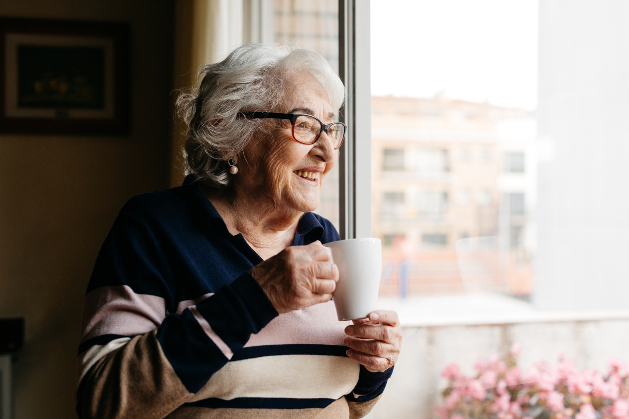 Portrait of an elderly woman holding a cup of coffee and smiling warmly while looking out a window.